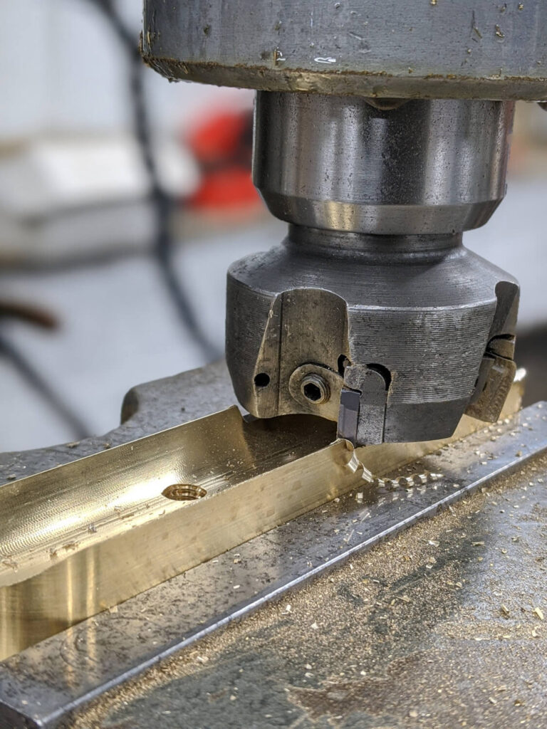 A close-up of a milling machine cutting a piece of metal, producing small shavings. The metallic surface and tool are clearly visible, highlighting the precision of the machining process.