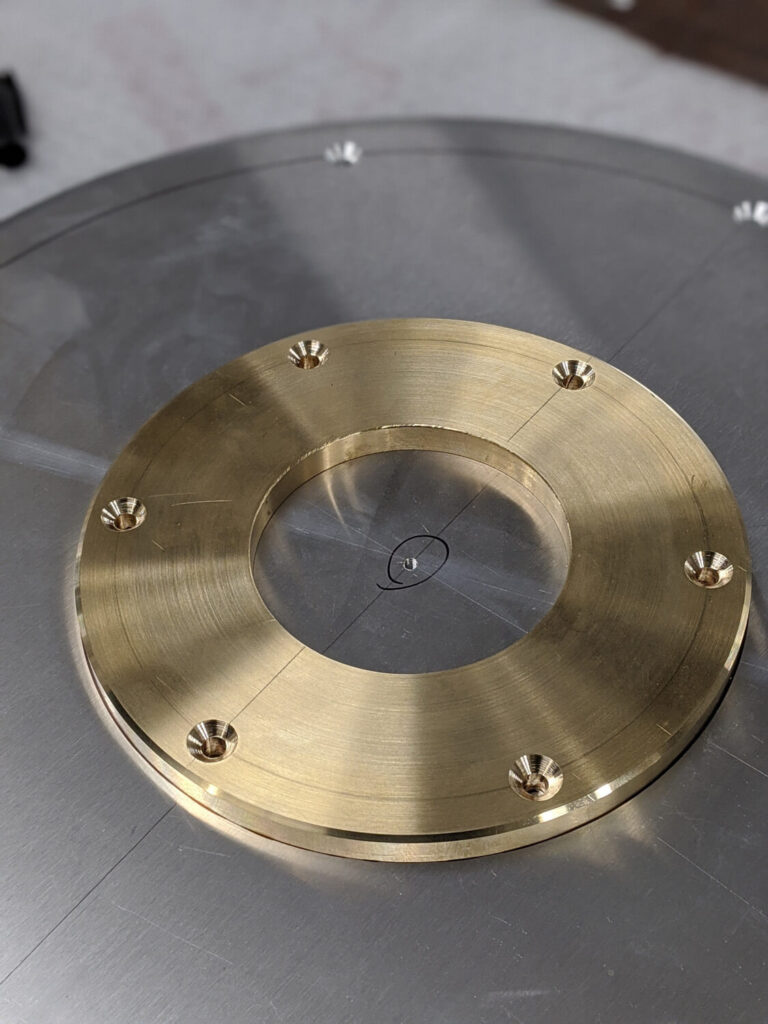 Close-up of a metallic circular object with a brass ring and several evenly spaced screws set on a reflective surface. The brass component is centered within a larger circular grey metal base. The metal is polished and has a gleaming finish.