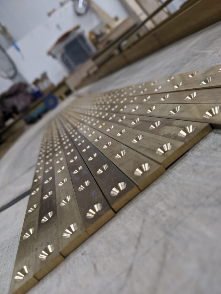 Close-up of metal strips with evenly spaced holes, laid out on a workshop table. The background shows blurred tools and equipment. The strips create a symmetrical pattern, emphasising precision and craftsmanship.