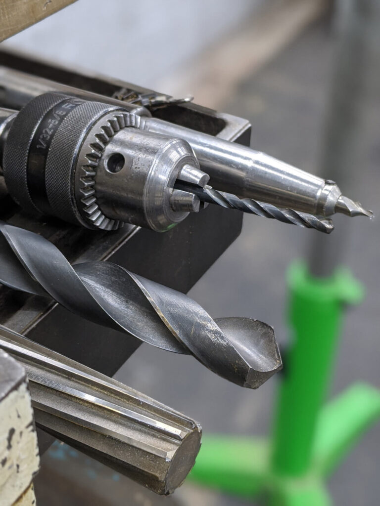 Close-up of two drill bits on a metal bench, with a blurred green base in the background. The larger drill bit is positioned horizontally in front of a smaller, vertical bit held in a chuck. The image showcases industrial tools.