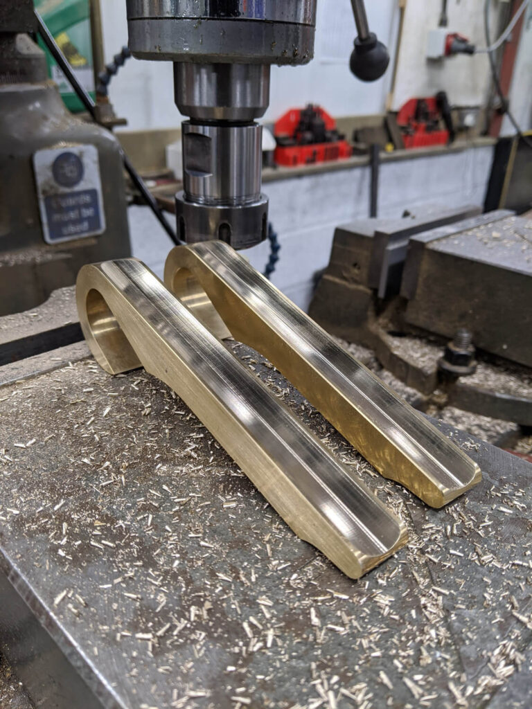 Close-up of a metal milling machine in a workshop with two brass components being worked on. Metal shavings cover the surface around the pieces. Tools and equipment are visible in the background.