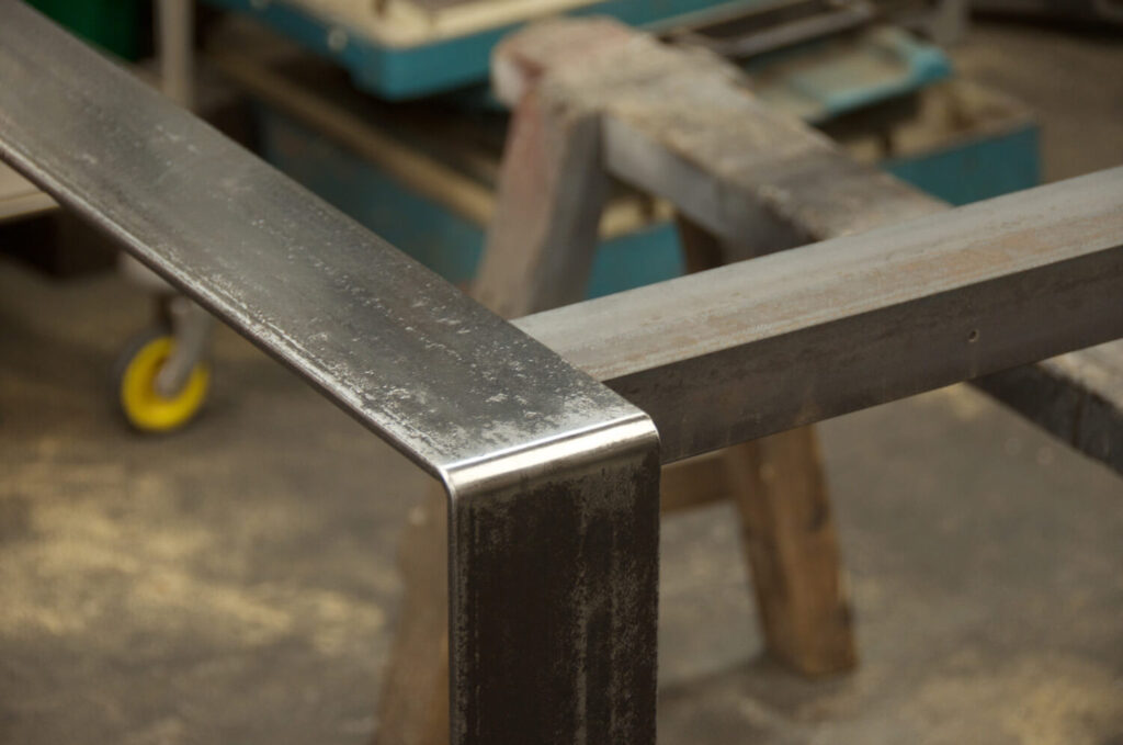 Close-up of a steel rectangular table frame joint in a workshop. The shiny corners contrast with the matte finish of the bars. Tools and equipment are blurred in the background, including a yellow-wheeled object.