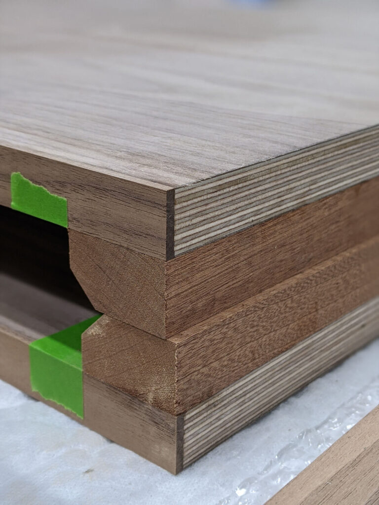 Close-up of a wooden table edge showing layered construction with exposed plywood and solid wood. Green tape is applied to some sections. The table surface has a light wood grain finish, while the sides are in darker tones.