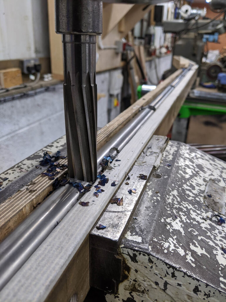 Close-up of a drill bit cutting into a metal surface, creating shavings. The equipment is set in a workshop, surrounded by various tools and machinery, with a textured surface underneath the workpiece.