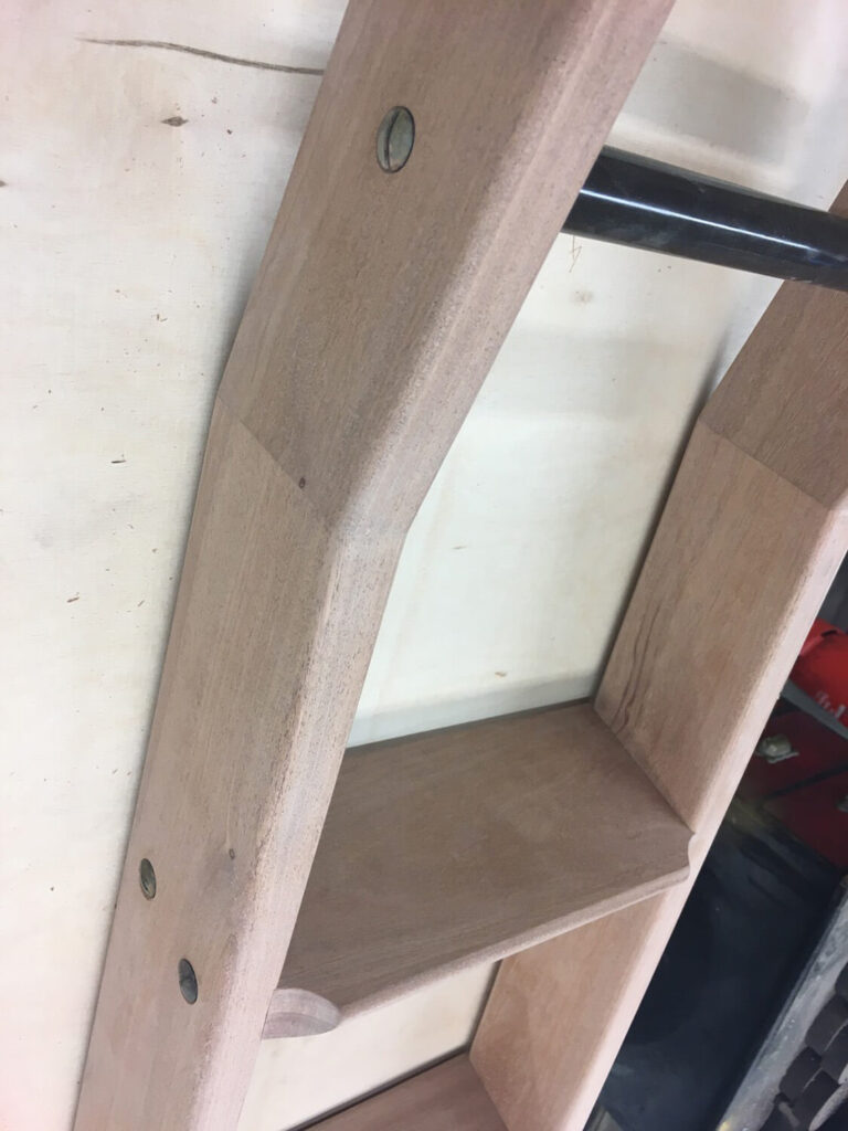 Close-up of a wooden library ladder leaning against a light-colored wall. The ladder has steel rivets and a smooth finish, with visible joints and steps. A portion of the rung hinge is visible.