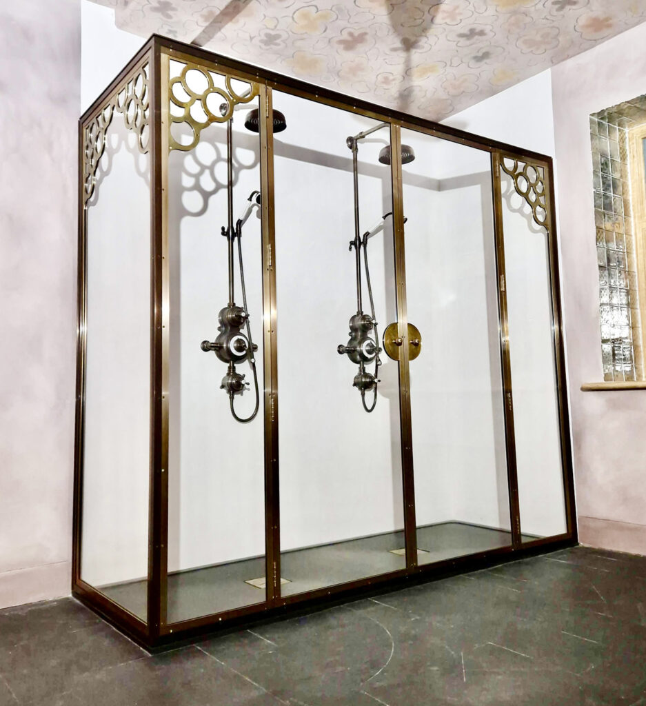 Ornate glass and brass shower enclosure with vintage-style shower heads and intricate pattern detailing on the frame, set against a tiled floor and a decorative ceiling.