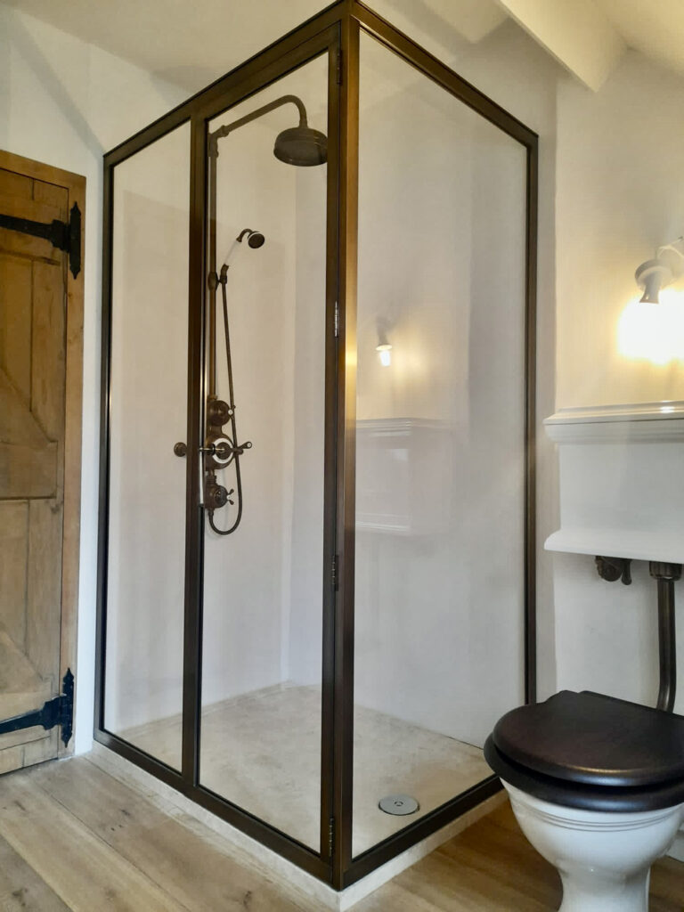 A bathroom features a corner glass shower enclosure with black metal frames and a rain shower head. There is a wooden door, a toilet with a dark seat, and a white wall-mounted sink beneath a light fixture. The floor is wooden.