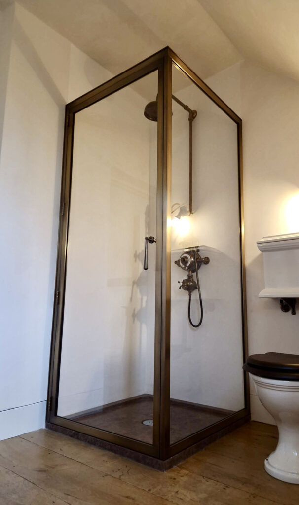 A bathroom with a glass shower enclosure featuring brass trim. Inside the shower are a showerhead, control valve, and handheld sprayer. Adjacent to the shower is a white toilet with a black seat, on a wooden floor.