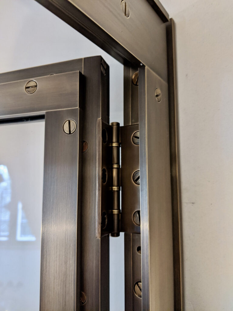 Close-up of a brass shower door hinge attached to a shower door frame. The hinge is brass with multiple screws fixing it to the frame. The wall is visible in the background, and part of a window reflection can be seen on the left.