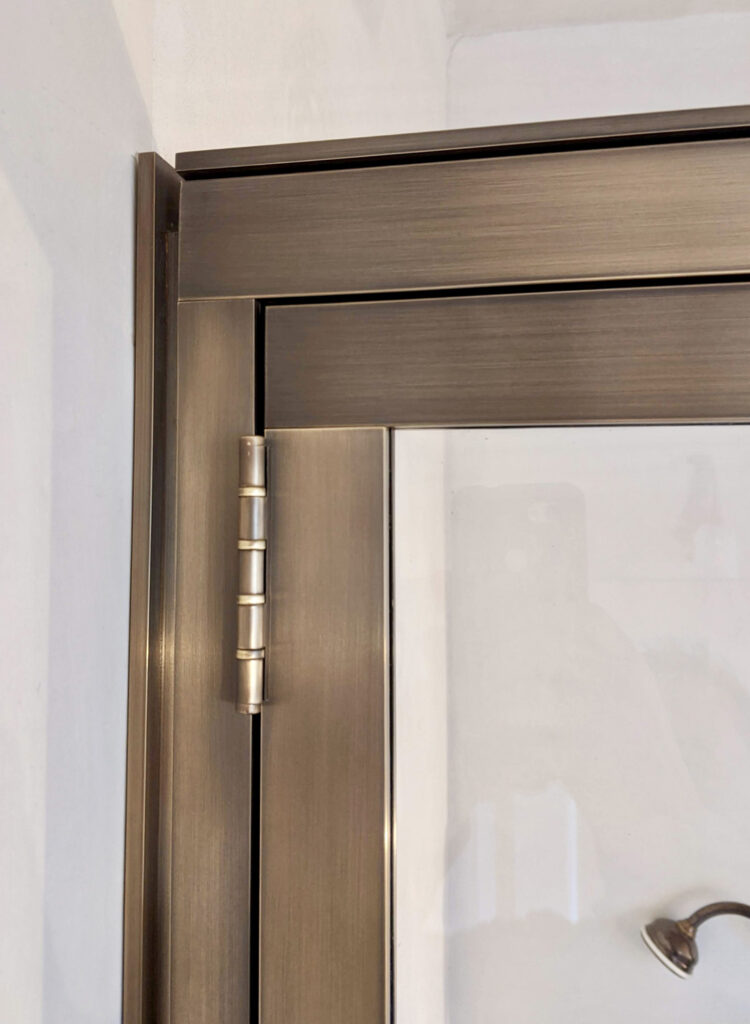 Close-up of a brass door frame showing a brass hinge. The hinge is attached to a shower door and frame, both featuring a brushed metal finish. The hinge has three cylindrical sections allowing the door to pivot.