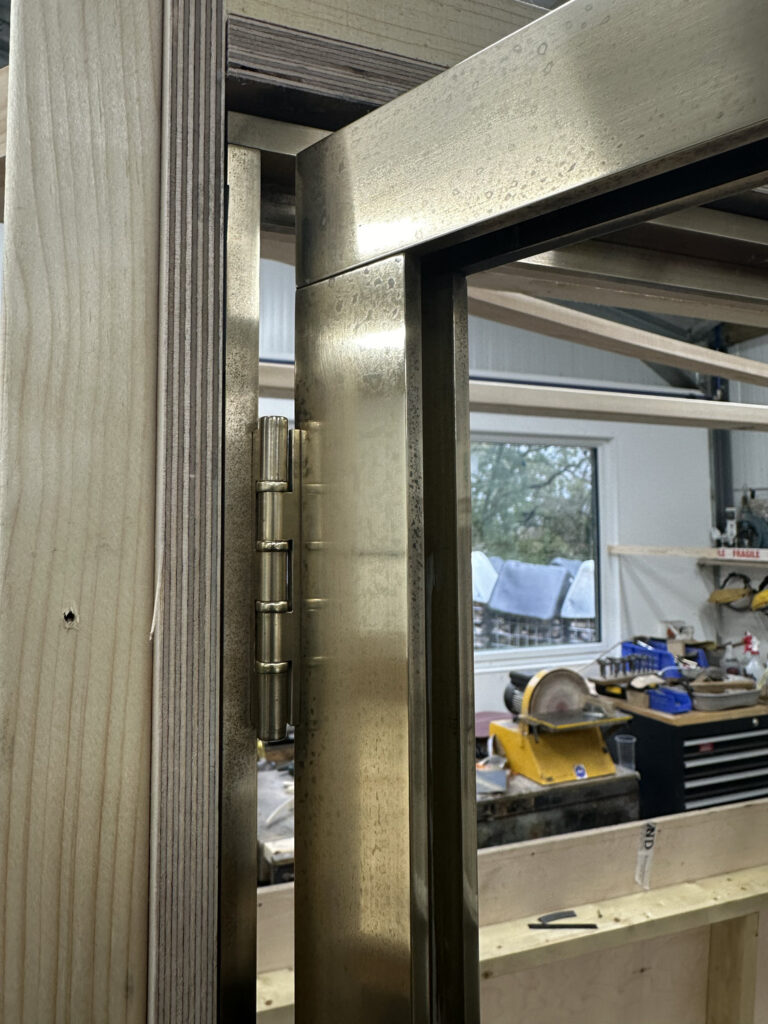 Close-up of a metal hinge on a partially open wooden door frame. The background shows a workshop with various tools, including a tool chest and a workbench near a window.