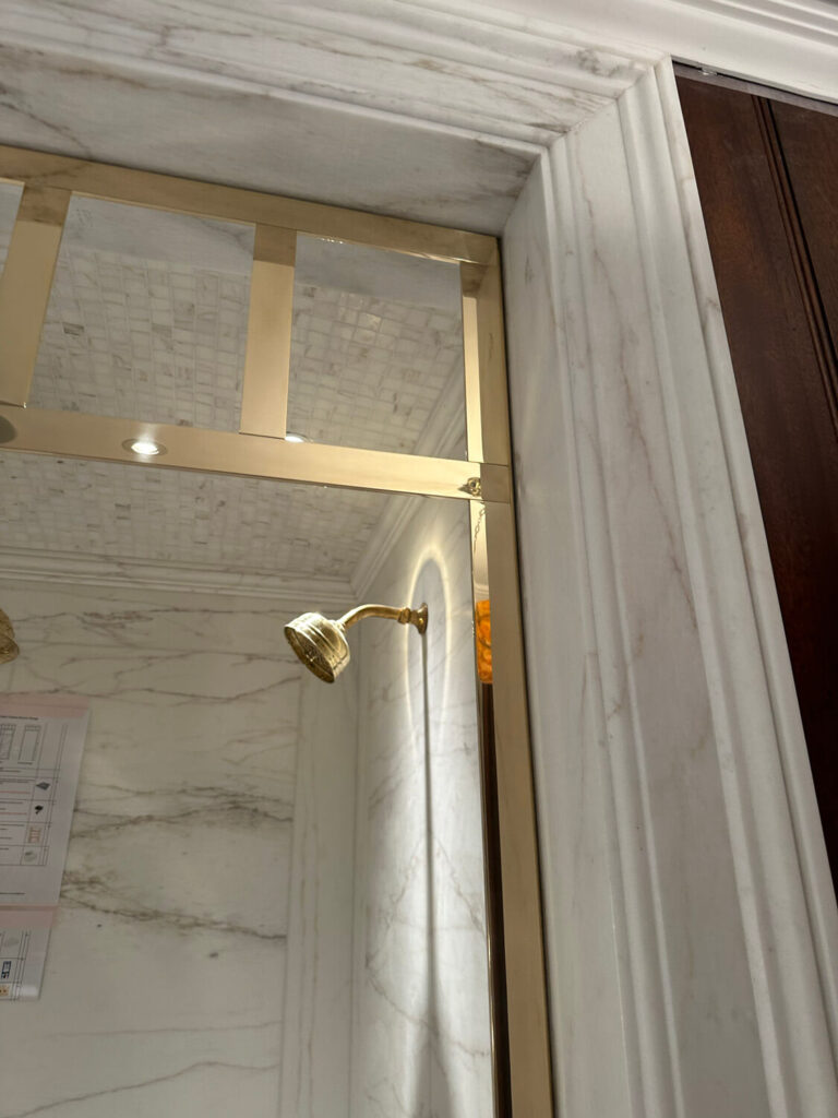 Elegant shower enclosure with marble walls and a glass door featuring gold trim. A brass shower head is visible inside, with a light reflecting off the surfaces. Wood paneling is on the right side of the image.