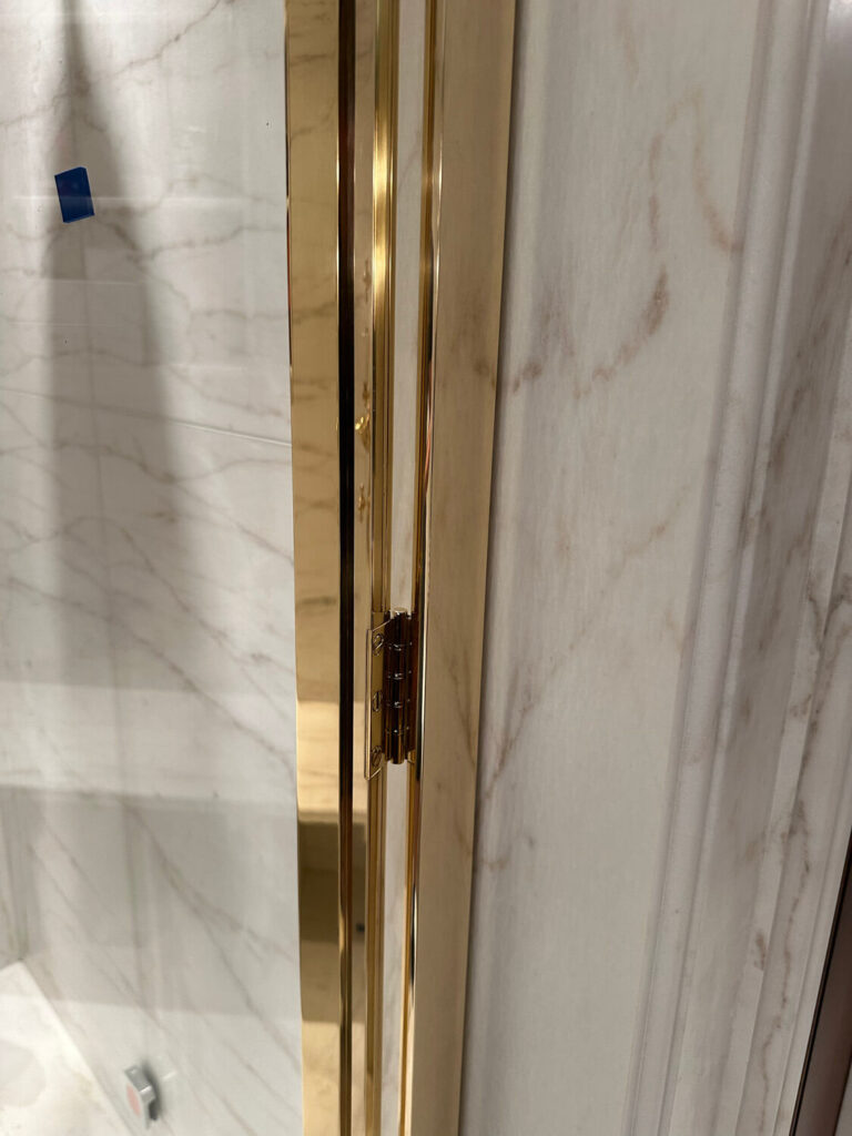 Close-up view of a bathroom shower bespoke brass shower screen. The walls feature a light marble pattern with subtle brown veining. The hinge of the door is visible, along with a small blue tape square on the wall.