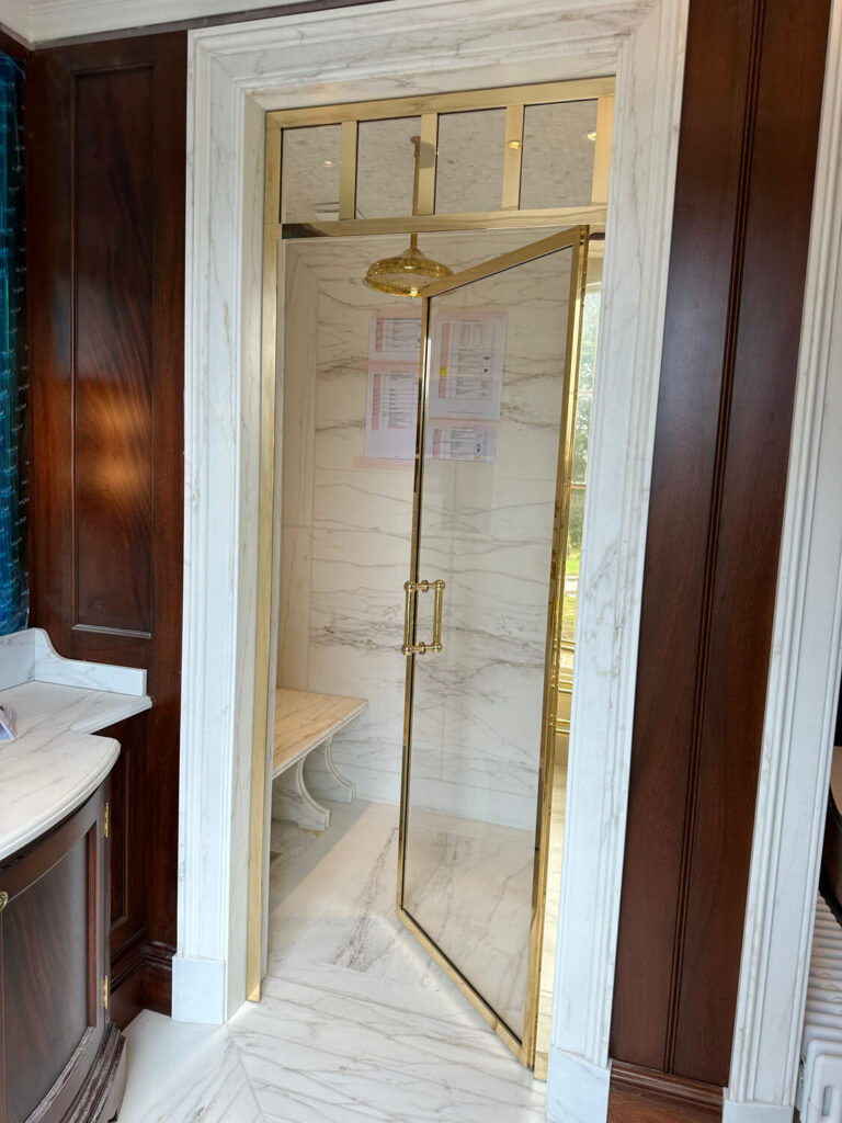 Luxurious bathroom featuring a marbled shower with a glass door and bespoke shower screen. The shower interior includes a built-in bench and brass showerhead. Rich wood panels and marble flooring complement the sophisticated design.