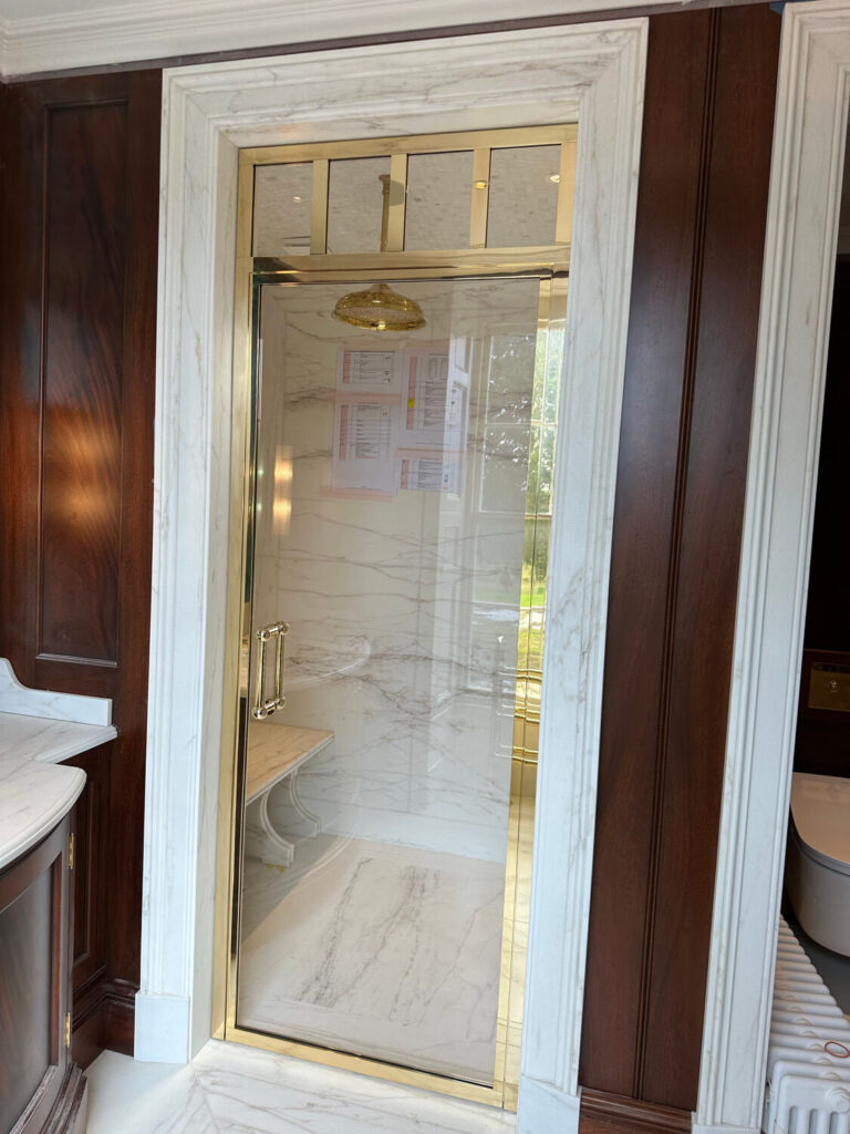 A luxurious shower with white marble walls and gold accents. It has a bespoke brass shower door with a brass handle and a rain shower head. Dark wooden panels surround the entrance, and theres a bench inside the shower.