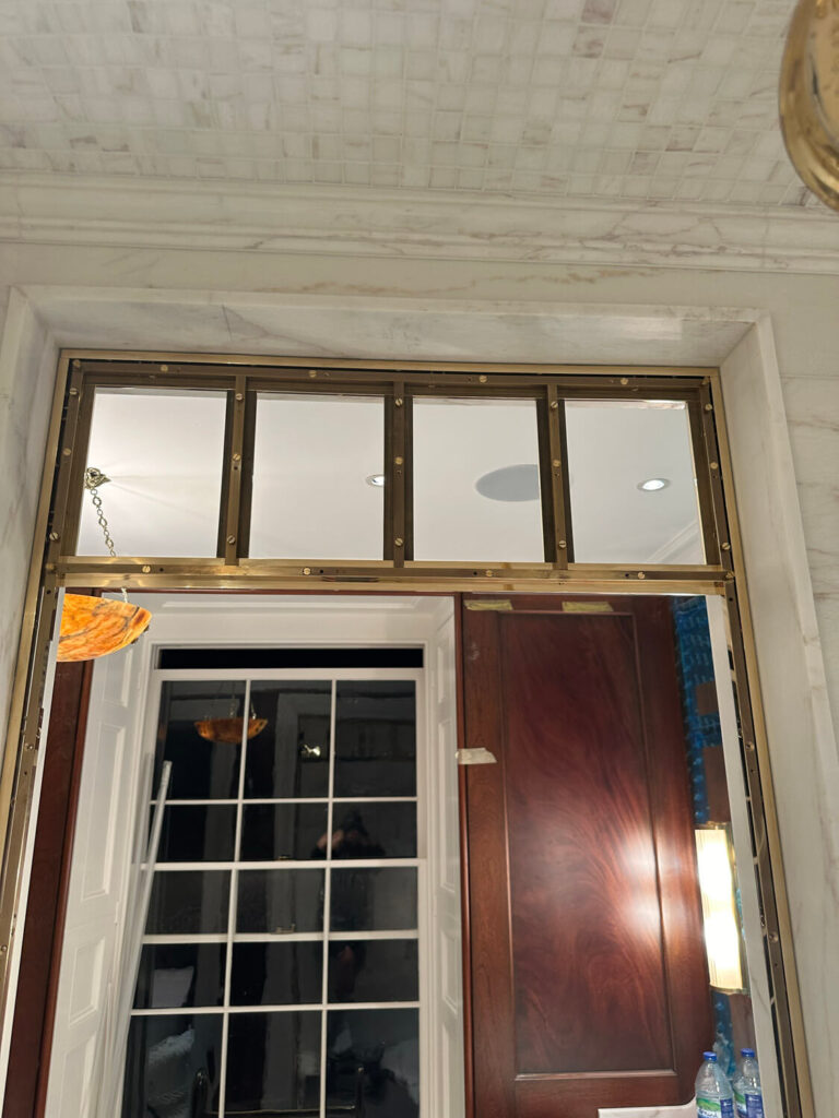 Interior scene with a marble wall and a window featuring a wooden frame with four glass panes. A glass door opens to reveal a glimpse of a lit room. Subtle ceiling lights illuminate the area, with a decorative pendant light partially visible.