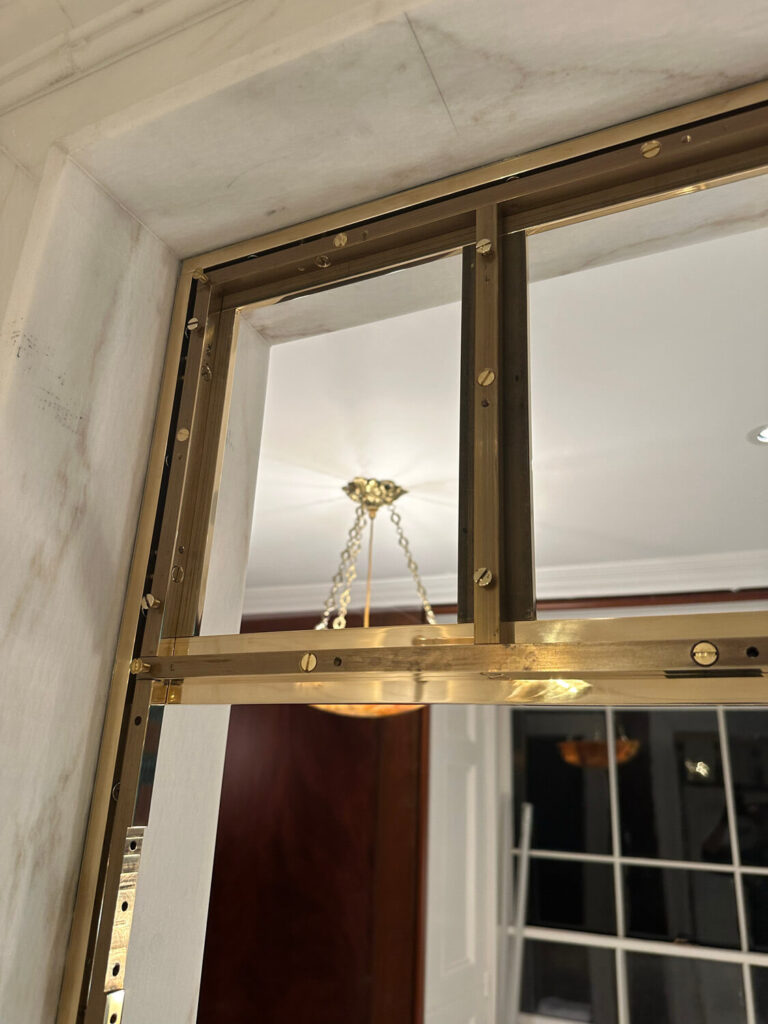 A gold metal frame with screws is attached to a marble wall. A chandelier with a frosted glass shade and chain is visible in the background, hanging from a white ceiling. The setting appears to be a luxurious indoor space.