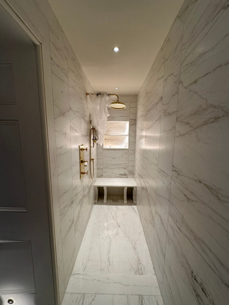 A narrow, elegant shower room featuring marble walls and floor. It has a gold rainfall showerhead, a small bench, and a window at the far end. Subtle recessed lighting illuminates the sleek design.