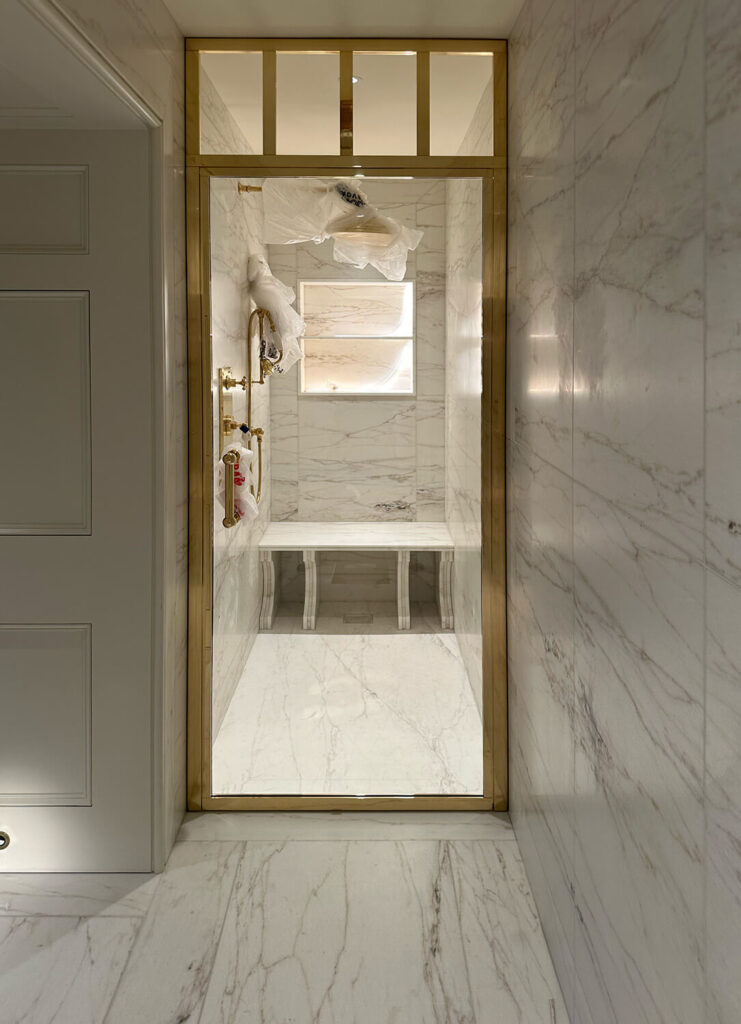 Luxurious marble shower with gold-framed glass door. Walls and floor feature white marble with gray veining. Inside, gold shower fixtures and a small window provide natural light. A towel rack and toiletries are visible on the left.