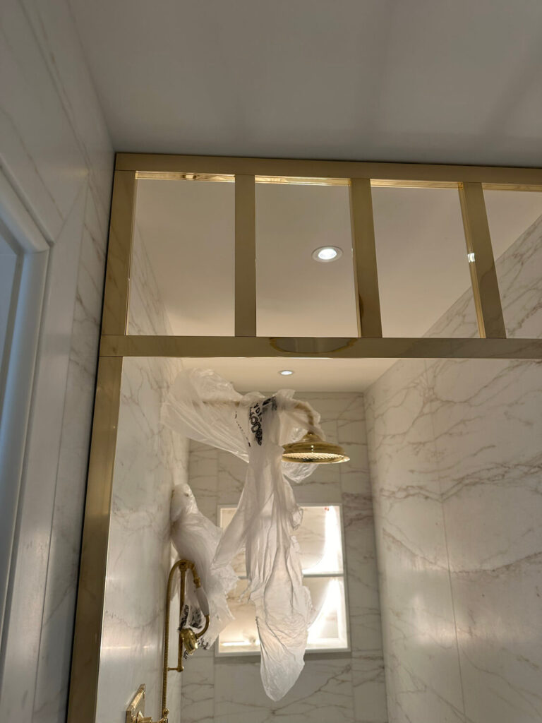 A shower with a gold-framed glass enclosure and marble tiles. A translucent plastic sheet is draped over the showerhead, with natural light coming through a small window above.