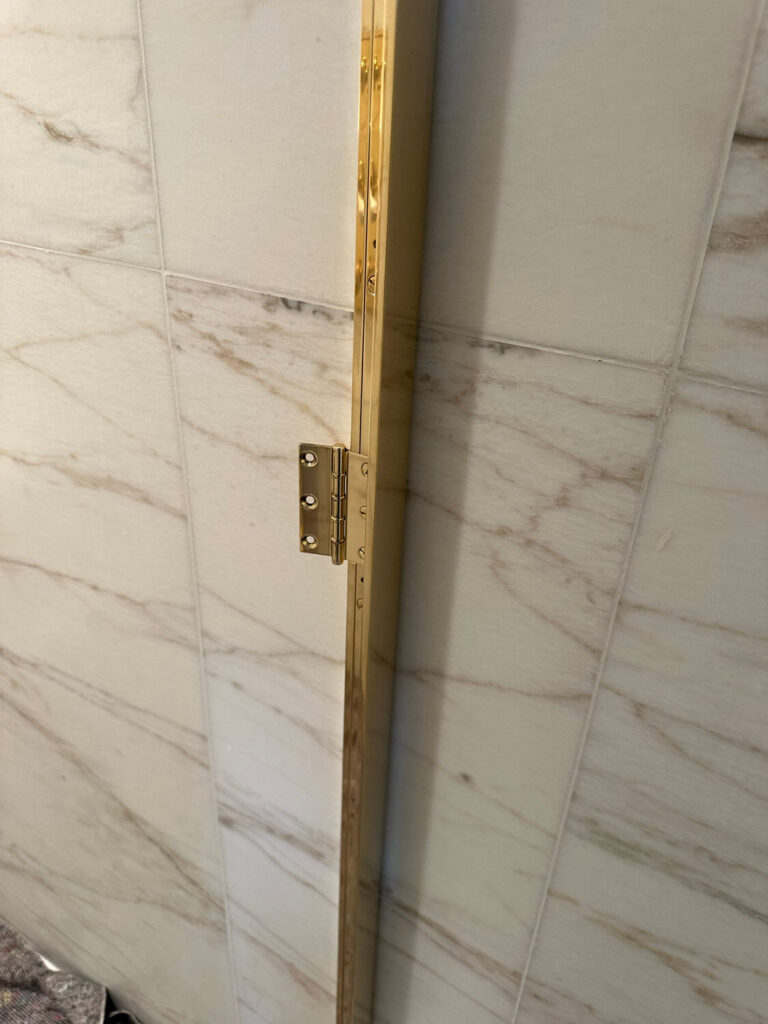 Close-up of a gold vertical metal strip with attached hinge against a wall of large, light-colored marble tiles with subtle gray veins. The strip is part of a door or panel fixture.