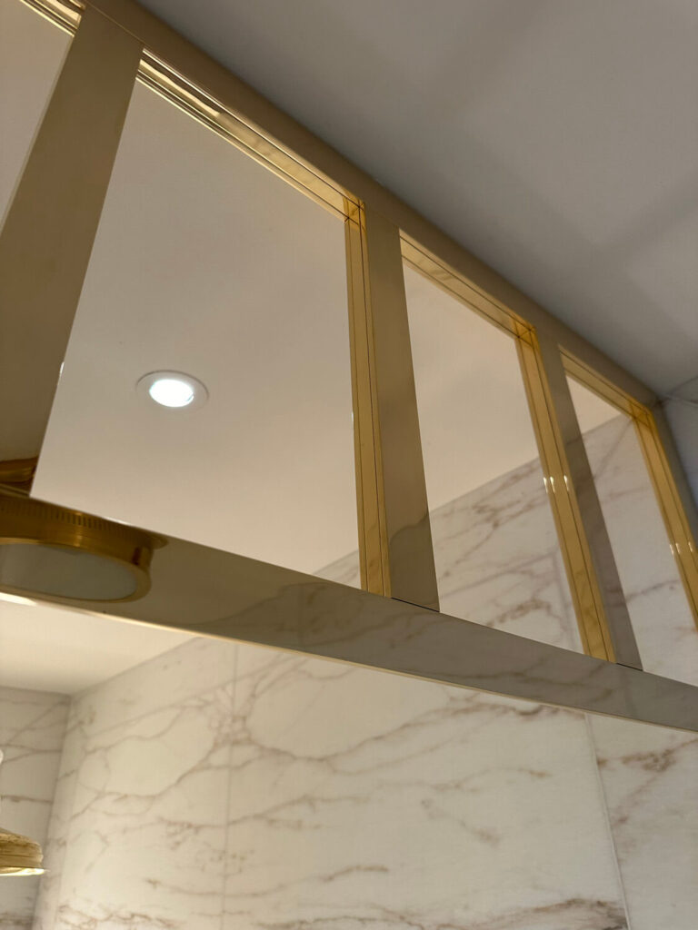 Close-up of an elegant bathroom with a shiny brass shower partition and white marble walls, featuring subtle gray veining. A recessed ceiling light illuminates the space, enhancing the luxurious ambiance.