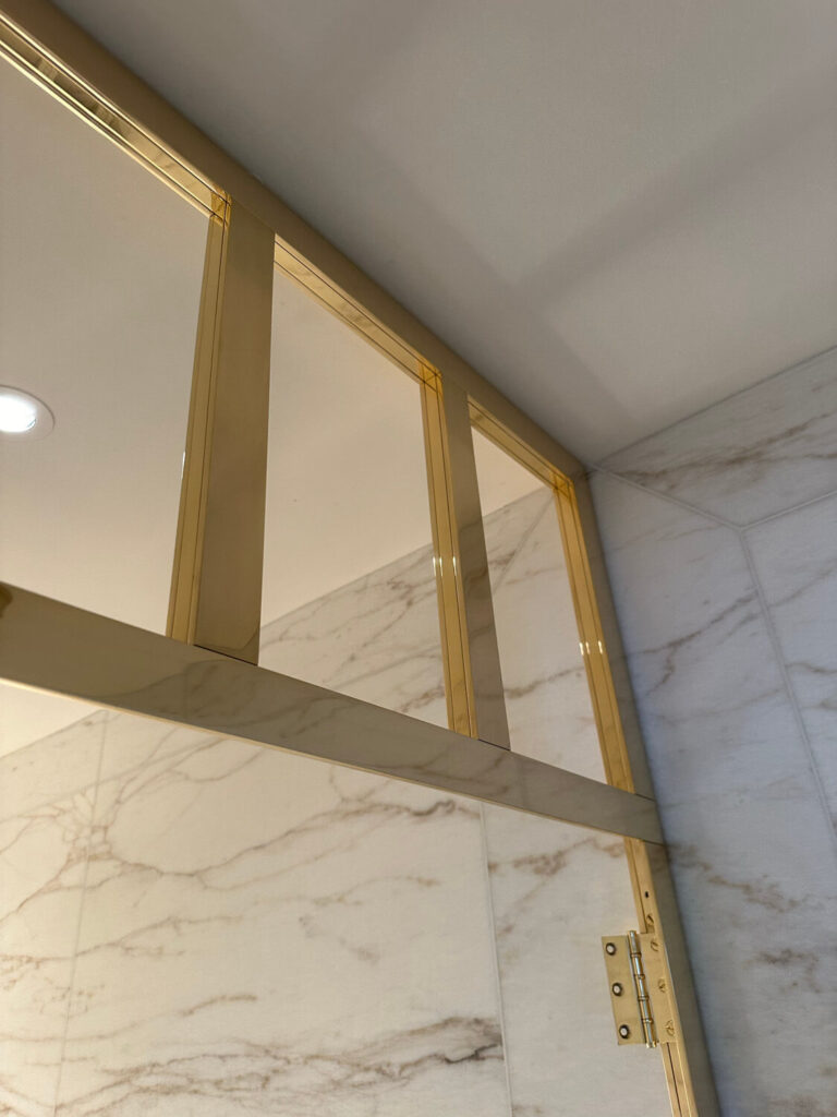 Close-up of an elegant shower enclosure with a gold metallic frame and hinge, contrasting against marble-patterned walls. The design features vertical glass panels and a seamless, modern aesthetic.