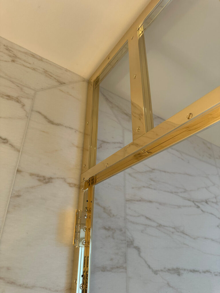 Marble-tiled shower with a corner of a glass door framed in shiny brass trim. The tiles have subtle veining, and the image captures a close-up of the doors hinge and framing against the marble.