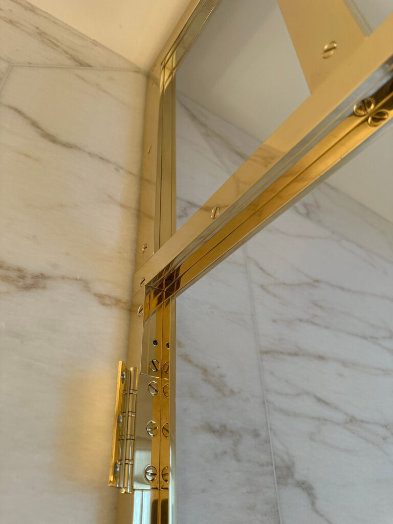 Close-up of a shiny gold-colored metal hinge attached to a glass panel, against a background of white, marble-like tiles. The hinge features decorative screws and a polished finish.