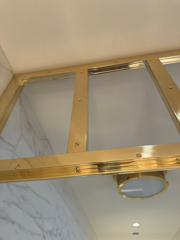 Close-up of a brass frame with glass panes near a ceiling, reflecting a ceiling light. The surrounding walls have a marble appearance with gray veining, creating an elegant, modern look.