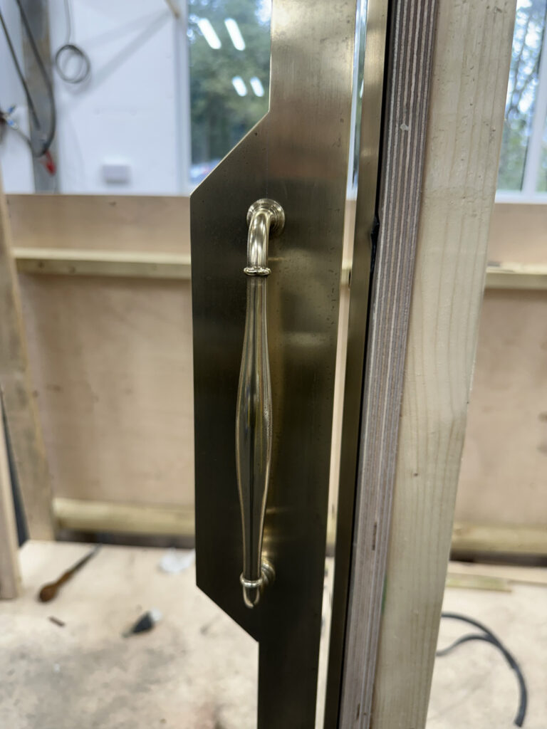 A close-up of a brass door handle mounted on a wooden door in a workshop. The door is partially open, revealing the unfinished interior space with tools and materials visible in the background.