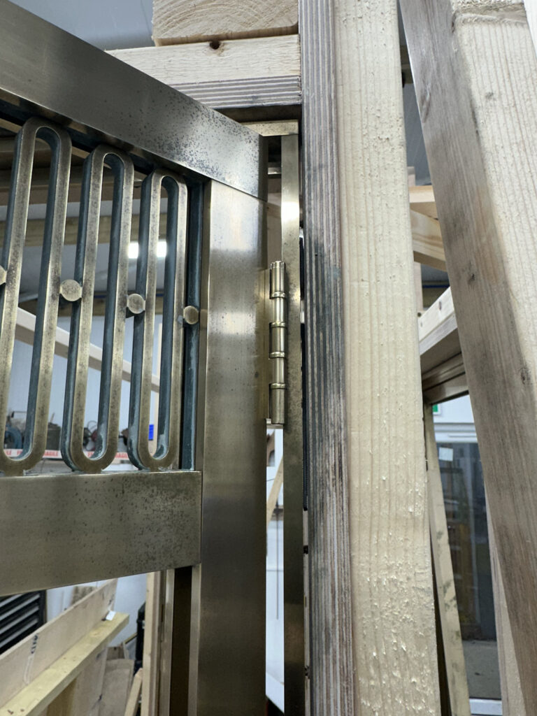 Metal gate with vertical bars is attached to a wooden structure using a sturdy hinge. The setup is inside a room with visible light fixtures, surrounded by unfinished wooden beams and planks.