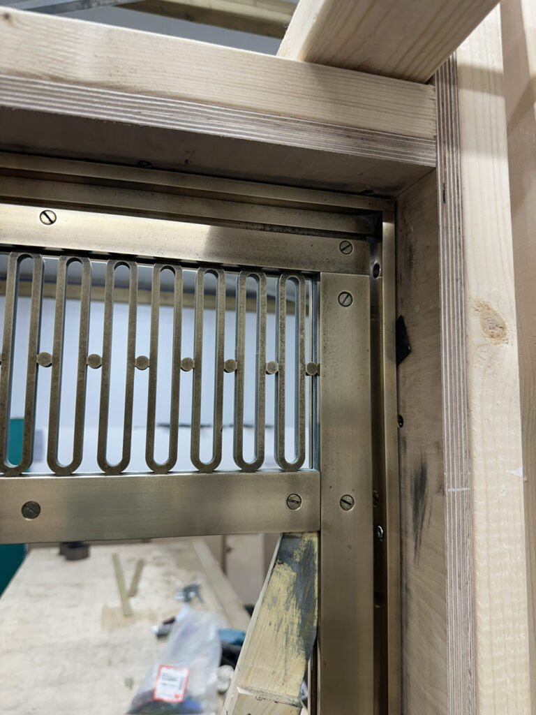 Close-up of a partially assembled wooden frame with a brass vent. The vent features vertical slots and is secured with screws. In the background, workshop tools and materials are visible on a table.