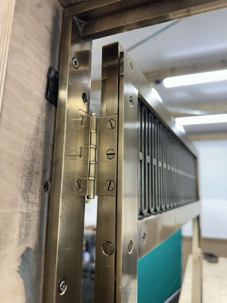 Close-up of a metal hinge on a door, showing intricate detailing with screws in a wooden frame. The hinge connects to a section with vertical metal bars and a teal panel, set in an industrial or workshop setting with bright lighting.