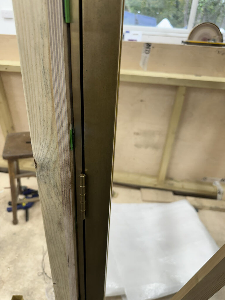 Close-up of a brass door hinge on a wooden frame with a view of a workshop interior. A stool and various tools are visible on the floor, and a wrapped item lies nearby. A window is in the background.