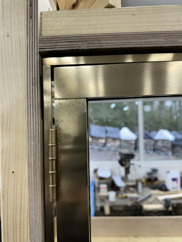 Close-up of a brass hinge on a wooden door frame, with the door partially open. The background shows a blurred view of a woodworking or construction workshop, including tools and equipment.